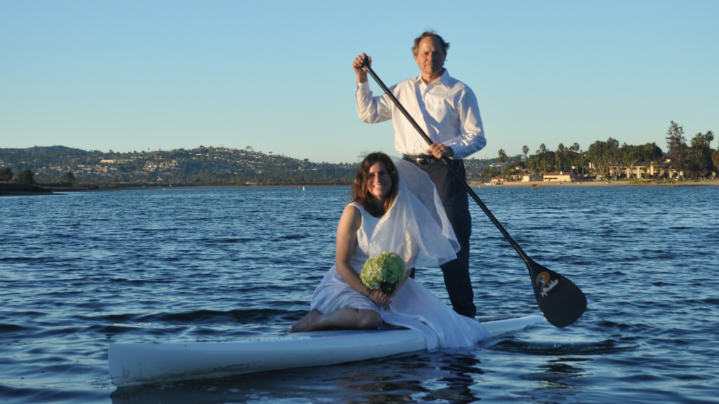 A man and woman in a wedding dress stand on a paddleboard on water
